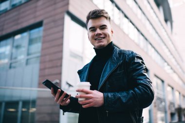 Deri ceketli, kahveli, cep telefonlu ve caddede modern tuğla bina karşısında kameraya bakan mutlu bir erkek.