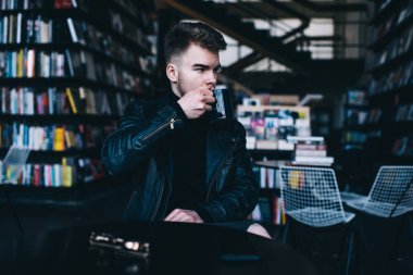 Yoğunlaşmış siyah deri ceketli genç adam modern kitapçıda masada otururken bir fincan sıcak içecek içiyor ve dalgın dalgın bakıyor.