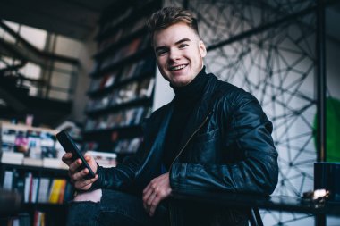 Yaratıcı kitapçıda sandalyede dinlenirken siyah günlük kıyafet giyen genç bir erkek cep telefonu kullanıyor.