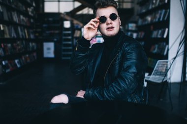 Ekin içeriği, modern ofisteki masada oturan siyah balıkçı yaka ceket ve güneş gözlüklü şık, tıraşsız genç adam.