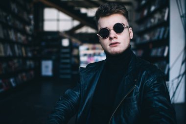 Güneş gözlüklü, balıkçı yaka ve siyah deri ceketli vahşi sakallı adamın portresi modern kafeteryada serinliyor arka planda kitap rafları var.