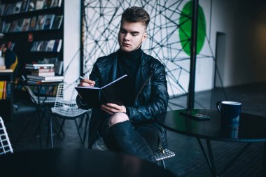 Düşünceli, genç, sakallı, modaya uygun, siyah kıyafetli, yaratıcı bir kafede çalışırken masada oturup notlar okuyan serbest bir erkek.