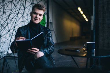 Gülümseyen stil sahibi, serbest çalışan, yırtık kot ve deri ceketli, yaratıcı çalışma alanındaki uzak iş sırasında organizatörden notlar alan şık bir adam.