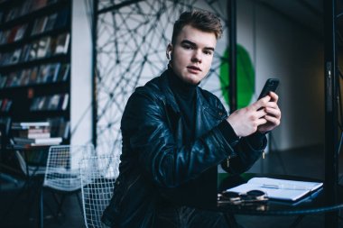 Moda deri ceketli, kablosuz kulaklıklı, cep telefonu kullanan ve kütüphanede açık not defteri ve güneş gözlüğüyle kameraya bakan genç bir hippi.