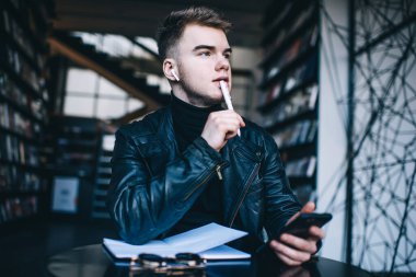 Kütüphanede açık not defteri ve cep telefonuyla otururken kablosuz kulaklık takan ve kalem ısıran genç bir erkek öğrenci.