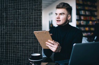 Siyah kazaklı, düşünceli, genç, sakallı, serbest bir erkek. Elinde bir not panosu, dizüstü bilgisayarında çalışıyor ve kahve içiyor.