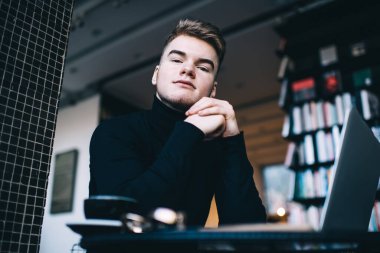 Düşük açılı, günlük kıyafetli, dizüstü bilgisayarlı bir erkek, masada oturuyor ve çağdaş iş yerinde bir fincan kahveyle rahatlıyor ve kameraya bakıyor.