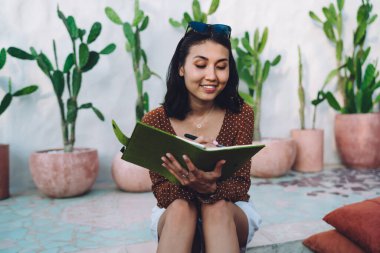 Neşeli, güler yüzlü, şık bir yaz elbisesi giyen genç bayan bahçedeki taş zeminde otururken günlüğe notlar yazıyor.