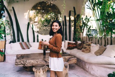 Moda kıyafetleri içinde Asyalı kadın serbest çalışan yazın tuvaletin yanındaki terasta durup kameraya bakarken cep telefonuyla çalışan