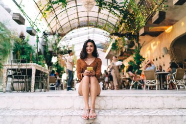 Zarif giyinen, merdivenlerde oturmuş, e-postaları kontrol eden, yaz tatilini çağdaş kafeteryada geçiren bir kadının düşük açılı görüntüsü.