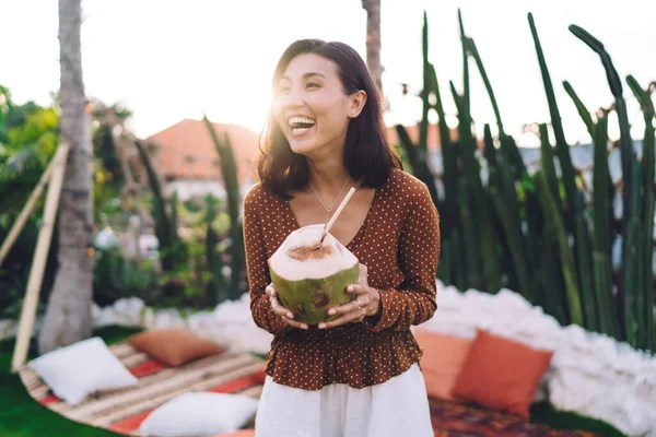 Polkaドットブラウスの若い民族の女性は陽気に笑って 日当たりの良いテラスに立っている間緑のシェルから新鮮なココナッツドリンクを楽しんで — ストック写真