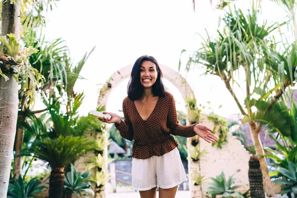 熱帯の裏庭にあるレンガ造りのアーチの前に立つ快適な光の服に身を包んだ若い民族の女性を笑顔とカメラを見ながらスマートフォンサーフィン — ストック写真