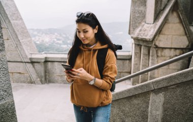 Gündelik kıyafet giyen Asyalı kadın turist elinde cep telefonu ile gezintiye çıkarken internet haritasında yerini kontrol ediyor, gezgin kadın akıllı telefon ve 4G bağlantısı kullanarak gideceği yeri keşfediyor