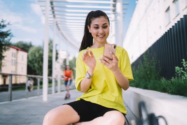 Sarı tişörtlü ve siyah şortlu mutlu genç Asyalı kadın yazın şehir caddesinde bankta otururken gülümsüyor ve mobil kullanıyor.