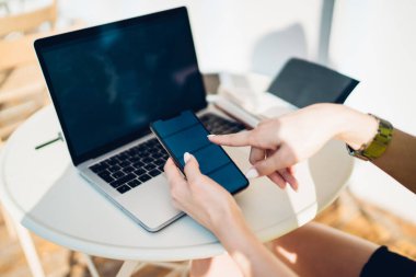 Kol saatindeki isimsiz bir kadın açık havada güneşli bir yaz günü uzaktan çalışma alanında dizüstü bilgisayarla proje üzerinde çalışırken haber okumak için telefonuna bakıyor.
