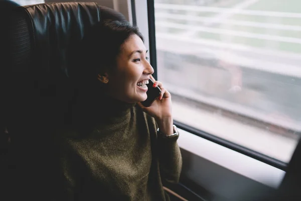 Cheerful asian female traveler laughing during mobile phone conversation using roaming connection on trip transporting via train,happy hipster girl making smartphone call sitting near window traveling