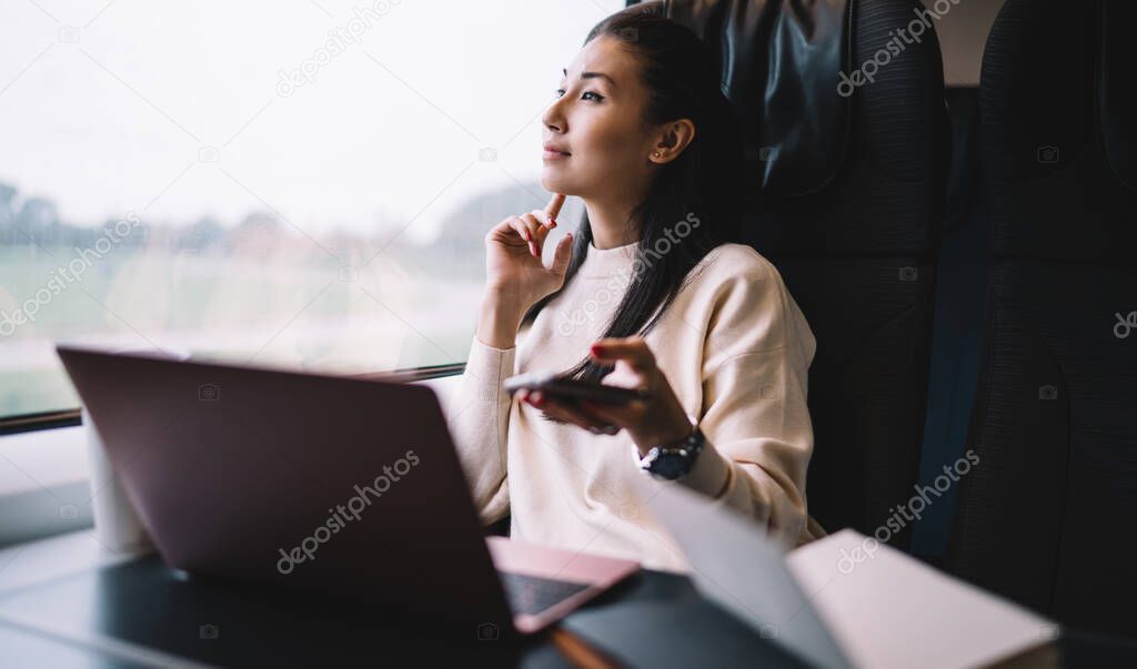 Pensive asian female blogger looking at window traveling by train thinking about publication for share in social networks, businesswoman with laptop computer sitting in wagon dreaming about vacation