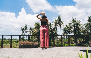 Avuçlarında telefonla bekleyen ve Bali 'de dinlenirken gözlerini kaçıran, şapkalı, tanınmayan kadın turistin arka görüntüsü.