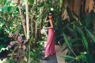 Elinde şapkayla tropikal verandada dikilmiş kameraya bakarken gülümseyen, gülümseyen zayıf bir kadın vücudu.