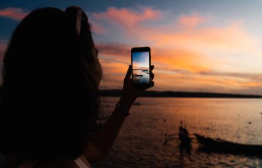 Tanımlanamayan bir kadın, deniz kenarında durup Bali 'deki deniz manzarasının fotoğrafını çekerken akıllı telefon kullanıyor.