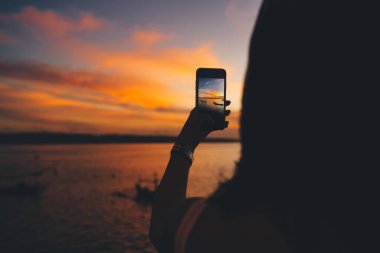 Gün batımında dalgalı denizin yanında duran ve gün batımını çekmek için akıllı telefon kullanan yüzü olmayan uzun saçlı bir kadının arka görüntüsü.