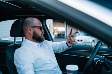 Optik gözlüklü kendine güvenen erkek sürücü, gelişmiş pahalı otomobil ve ulaşım konseptiyle otomatik pilotu test ederken akıllı telefona odaklanmış göz koruması sağlar.