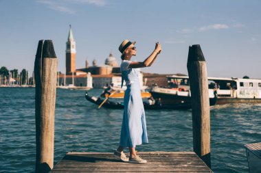 İtalya 'da Venedik rıhtımında güneş ışığında beklerken, şapkalı, gülümseyen ve cep telefonuyla selfie çeken neşeli genç bir kadın.