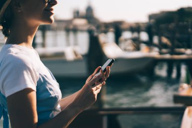 İtalya 'da Venedik rıhtımında dikilirken günlük giysiler içinde elinde cep telefonuyla bekleyen neşeli genç bayanın yan görüntüsü.