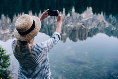 Huzurlu Karersee Gölü kıyısında dururken akıllı telefon kullanarak sıradağların fotoğrafını çeken genç bir kadın.