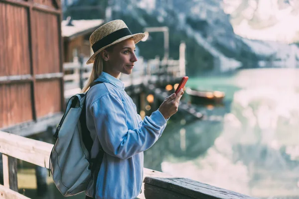 穿着休闲装和帽子站在湖边木制码头边 手持智能手机望着远方的年轻而积极的女游客的侧视图 — 图库照片