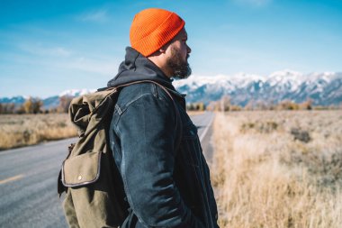 Sakallı, erkek gezgin, sıcak kıyafetler giymiş, sırt çantalı, yol kenarında duran kırsal kesime bakan.