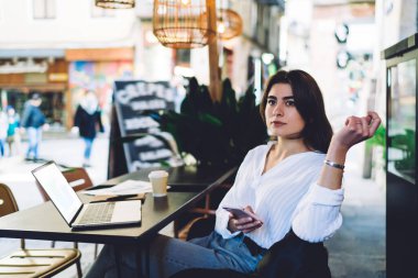 Elinde cep telefonu cihazıyla tefekkür eden özgür bir kadın sokak terasında boş zamanlarında düşünceli bakışlarını kaçırıyor, akıllı telefonlu beyaz blogcu yayın fikri üzerinde kafa yoruyor.