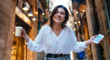 Kafein içeceği ve akıllı telefon cihazıyla turistik seyahat sırasında kameraya gülen, kulaklıklı neşeli bir kadının yarım boy portresi.