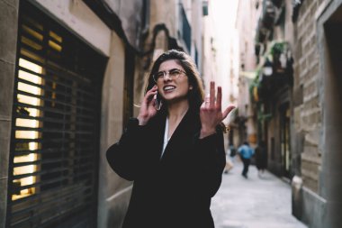 Resmi kıyafetler ve gözlükler içinde kafası karışmış bir kadın dar bir sokakta duruyor ve yürürken akıllı telefondan konuşuyor ve başka tarafa bakıyor.