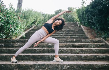 Beyaz tayt ve spor ayakkabılı, açık havada yoga yaparken Genişletilmiş Kenar Açısı pozu veren, etnik kökenli genç bir bayan.