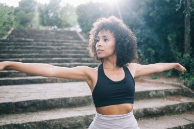 Kıvırcık saçlı, spor tişörtlü, kendine güvenen genç bir kadın güneşli bir günde yeşil parkta 2. Savaşçı pozu verirken gözlerini kaçırıp ellerini uzatıyor.