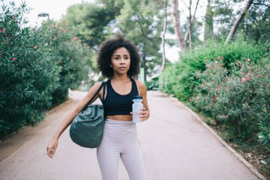Afro-Amerikan spor kıyafetleri içinde kendinden emin bir kadın ve bir şişe suyla parkta yürüyor ve düşünceli bir şekilde başını çeviriyor.