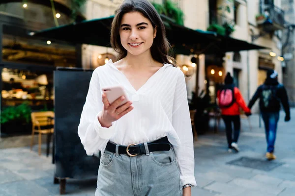 Happy stylish female in trendy outfit with toothy smile browsing smartphone while standing on pavement near modern cafe and looking at camera