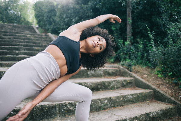 From below of crop happy young ethnic woman with curly hair standing in pose Extended Side Angle during practicing yoga asana on stairs in park