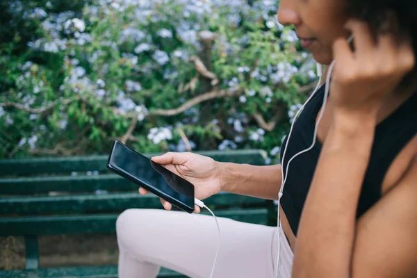 Crop black female in sportswear using smartphone with black screen for listening music in earphones while relaxing after training on bench in flowering park