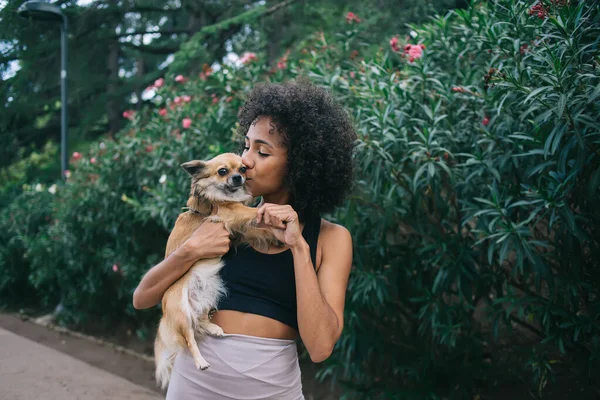 スポーツウェアの女性のコンテンツキスや腕の中で犬を保持しながら 市内通りの公園で茂みの近くに立って — ストック写真