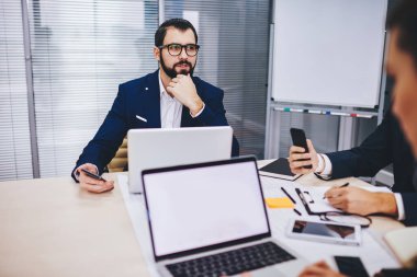 Optik gözlüklü, düşünceli bir erkek yönetici. Masa başındaki ortak çalışma sürecinde finansal araştırmalar üzerine kafa yoruyor. Laptop bilgisayarıyla, kopya alanı olan boş bir ağ defteri.