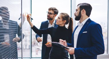 İş stratejisi planlamak için modern cam tahtaya şema çizen Kafkasyalı meslektaşların yan görünümü, ofis içi çalışma sürecinde deneyimli erkek ve kadın ortakların işbirliği