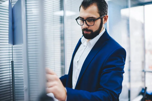 Confident bearded proud ceo in glasses making notes for financial reports, concentrated executive director dressed in formal wear writing benchmark information of working process standing in office