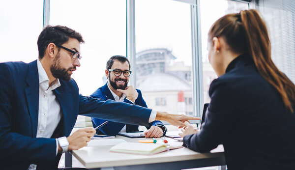 Portrait of cheerful male entrepreneur in optical eyewear smiling at camera while successful colleagues making laptop booking during collaborative meeting, Caucasian partners discussing startup