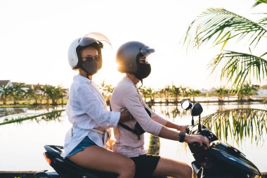 Kasklı ve maskeli, sakin göl kenarında motosiklet süren ve birlikte tropikal tatil köyünde vakit geçirirken gözlerini kaçıran tanınmamış genç çiftin yan görüntüsü.