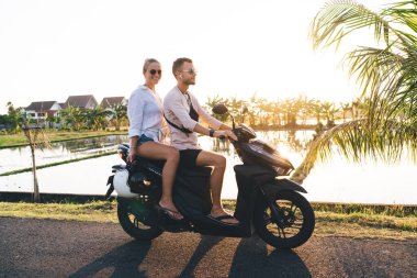 Gözlüklü mutlu çift sıcak kırsalda scooter kullanıyor ve tatil boyunca birlikte dinlenirken yaz günbatımının tadını çıkarıyor.