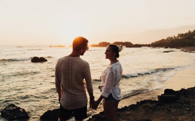 Gün batımında kumlu sahil kenarında duran tanınmayan bir kadın ve erkek akşam saatlerinde birbirlerine bakarken el ele tutuşuyorlar.