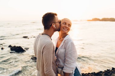 Güneş gözlüğü ve kablosuz kulaklıklı olumlu erkek. Gün batımında deniz kenarında dinlenirken kadının yanağından öpüyor.