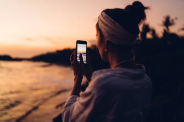 Saçında eşarp olan, ıslak zeminde dururken muhteşem günbatımının fotoğraflarını çeken anonim kadın blogcu.